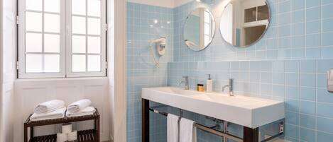 A bathroom with a white sink, two mirrors, a towel rack with towels, a hairdryer, and a window with blinds.