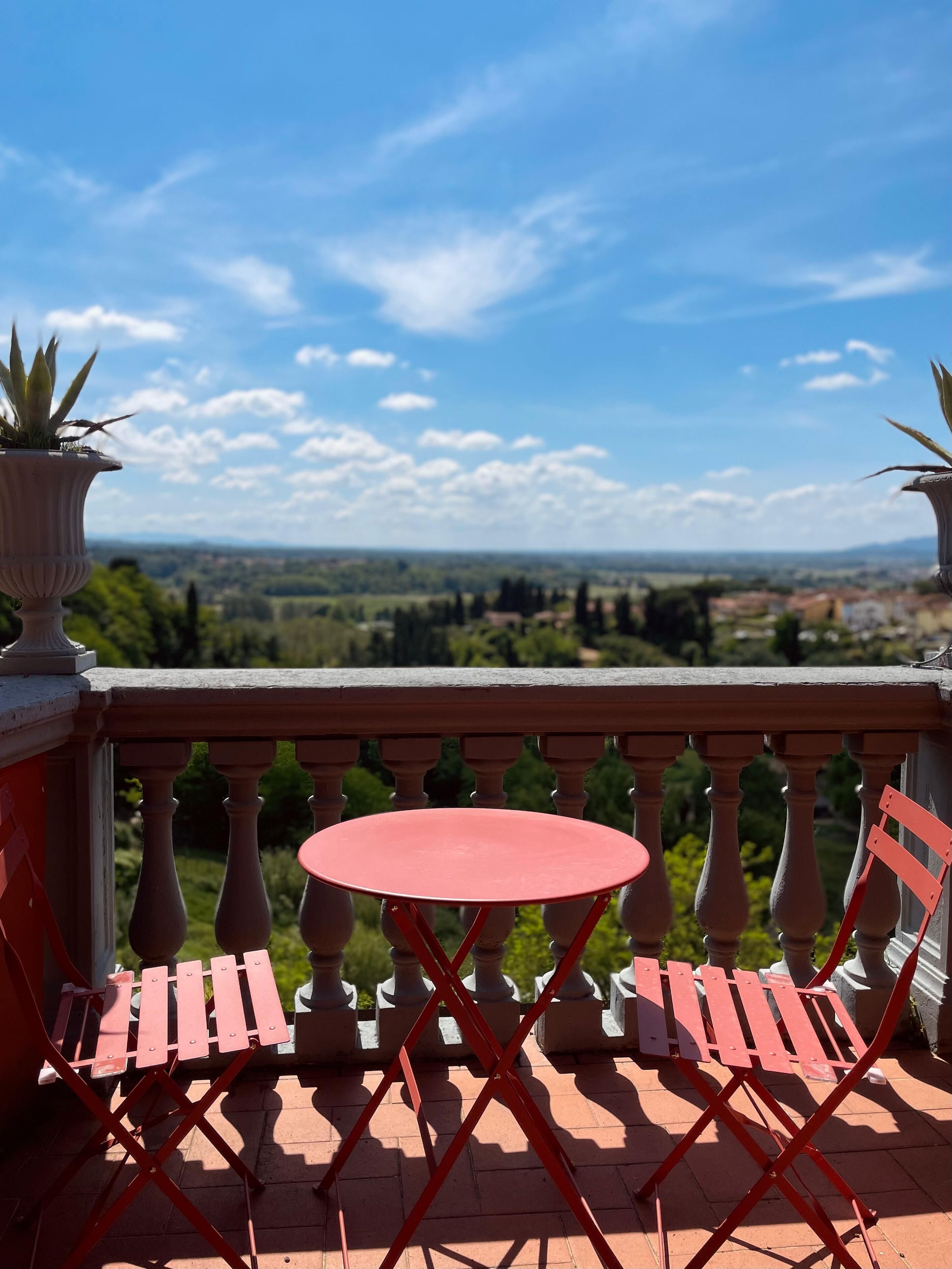 A balcony with a red table and chairs, overlooking a scenic landscape.