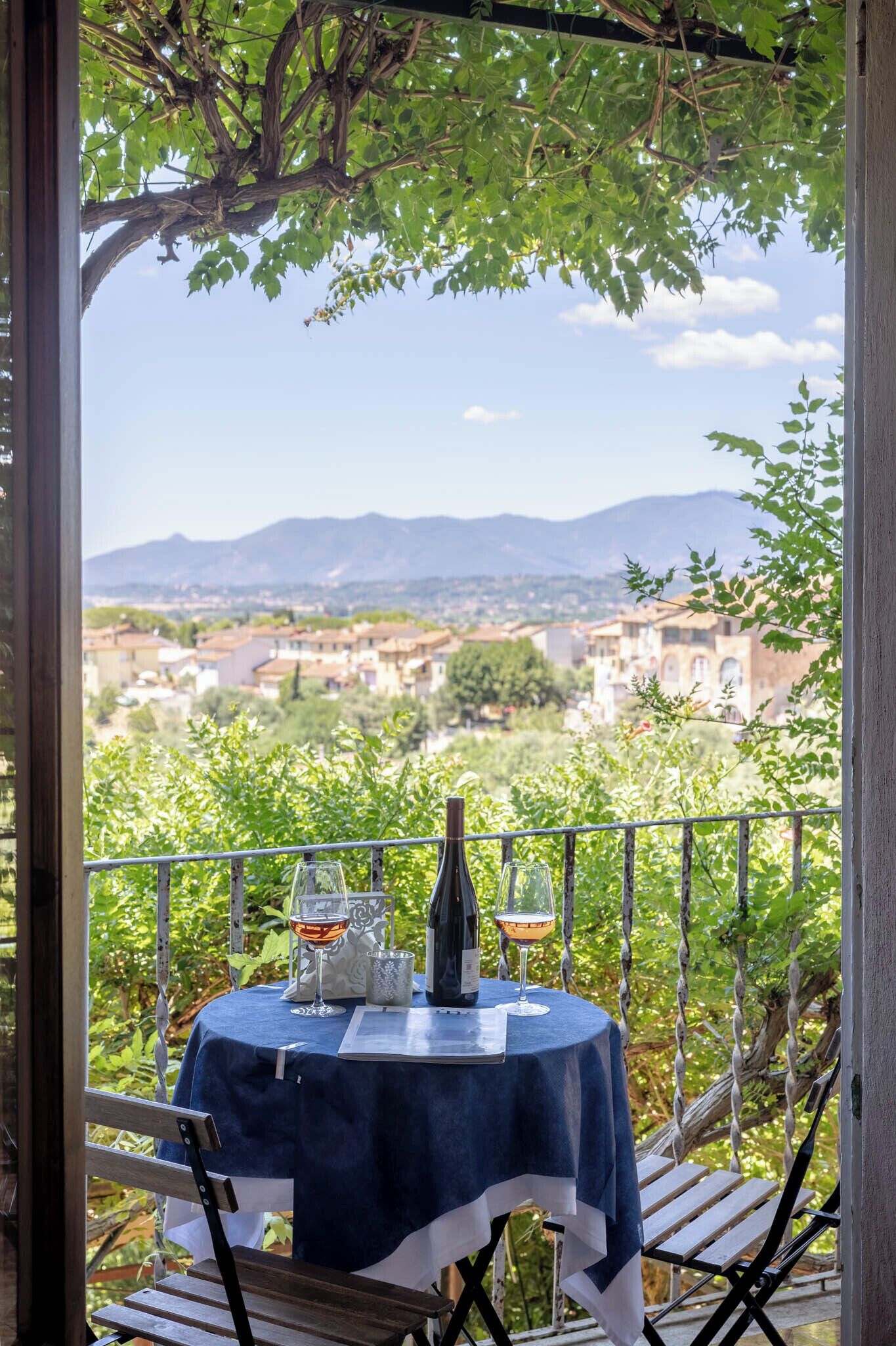 Un balcón con una mesa para dos, con vista a un paisaje pintoresco que incluye edificios y montañas.