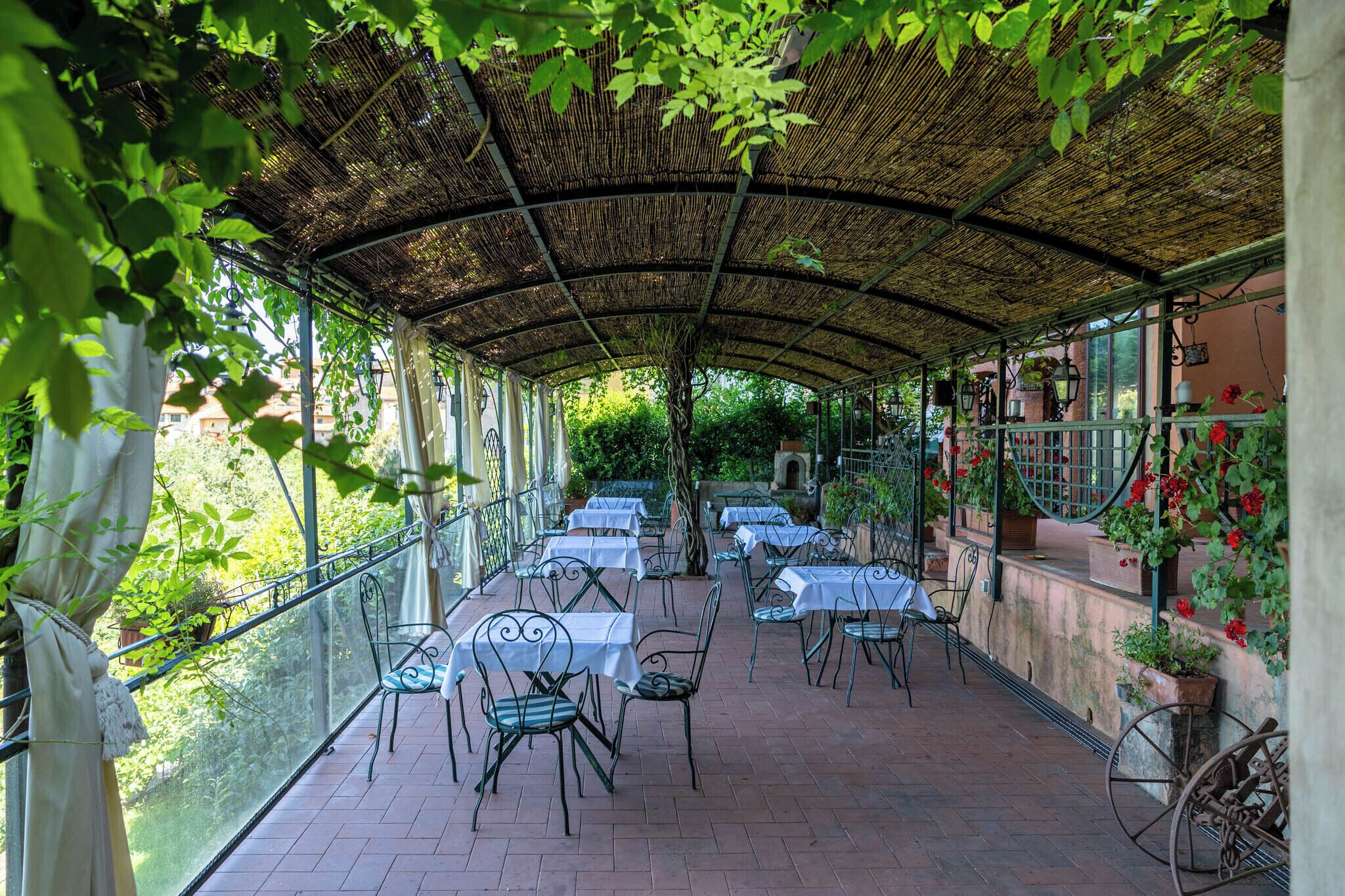 An outdoor patio with tables and chairs, covered by a woven roof, surrounded by greenery and potted plants.