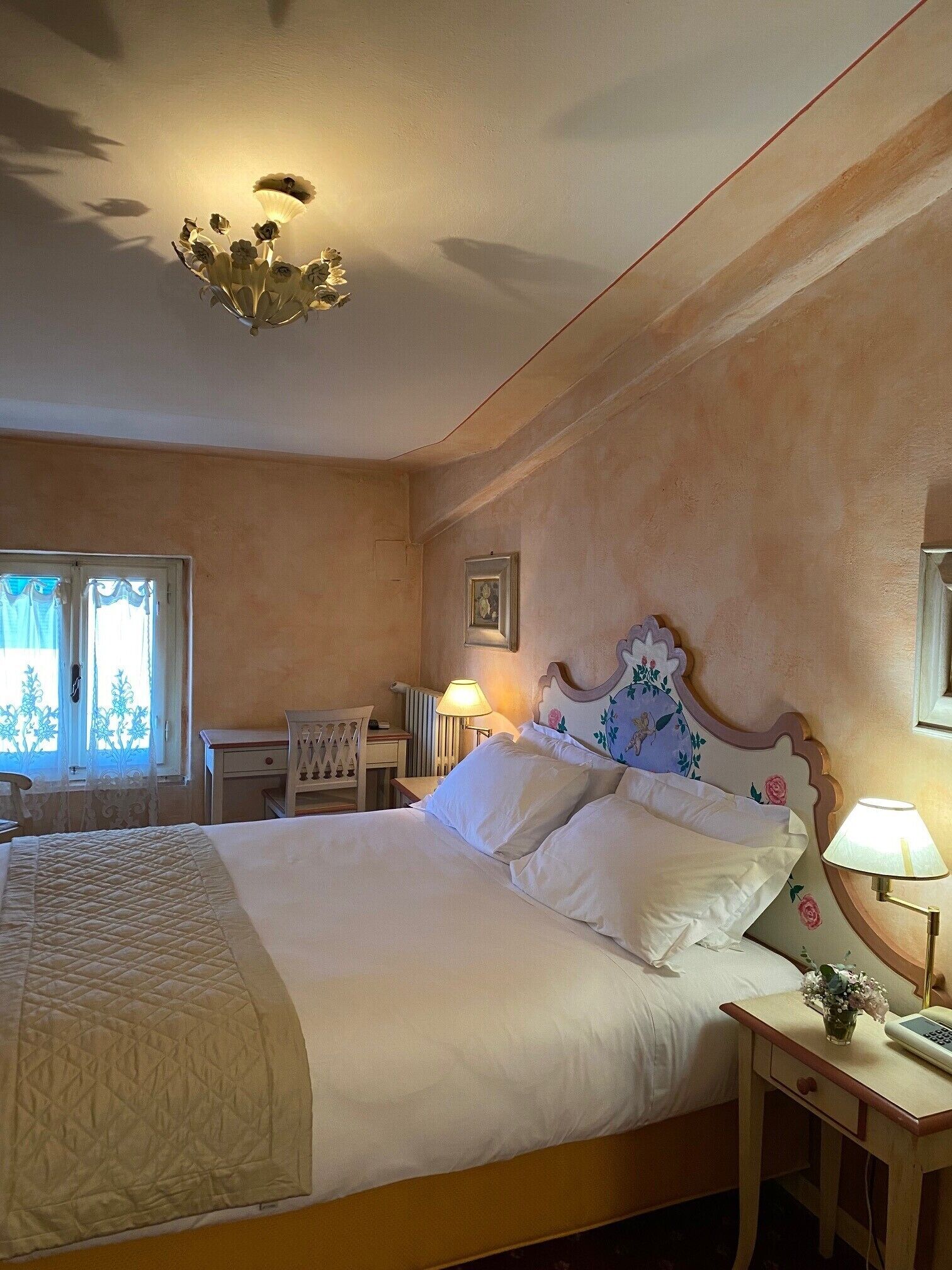 A bedroom with a large bed, a bedside table with a lamp, a chair, a desk, and a window with curtains.