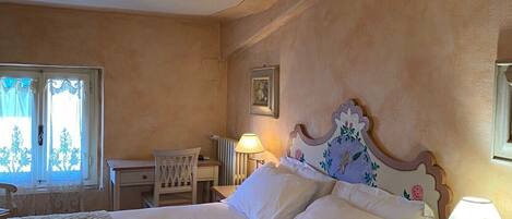 A bedroom with a large bed, a bedside table with a lamp, a chair, a desk, and a window with curtains.