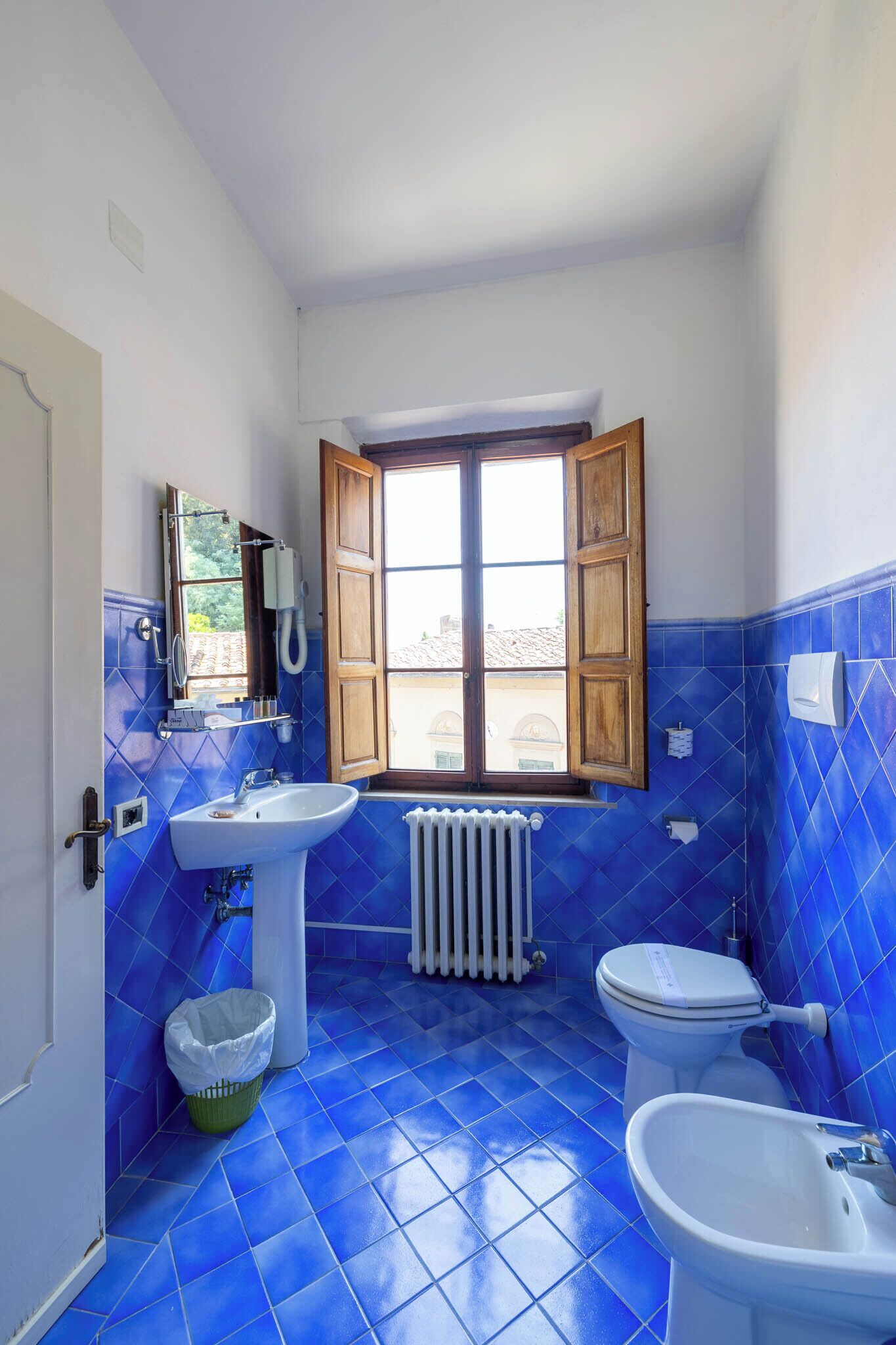 Un baño con azulejos azules, una ventana, un radiador, un lavabo, un inodoro y un espejo.