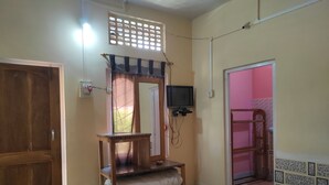 Une pièce avec une porte en bois, un lit, une petite table sur laquelle sont pliés des couvertures, un téléviseur fixé au mur et une fenêtre ornée d’un paravent décoratif.