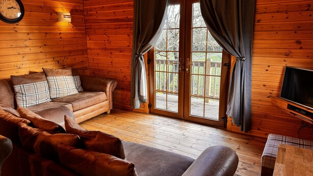 Un interior de cabaña acogedor, con detalles de madera, un sofá de cuero marrón, un televisor y un ventanal con cortinas.