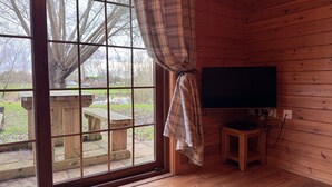 Une pièce de cabane en bois avec une grande fenêtre, une table en bois à l’extérieur et un téléviseur posé sur un meuble.