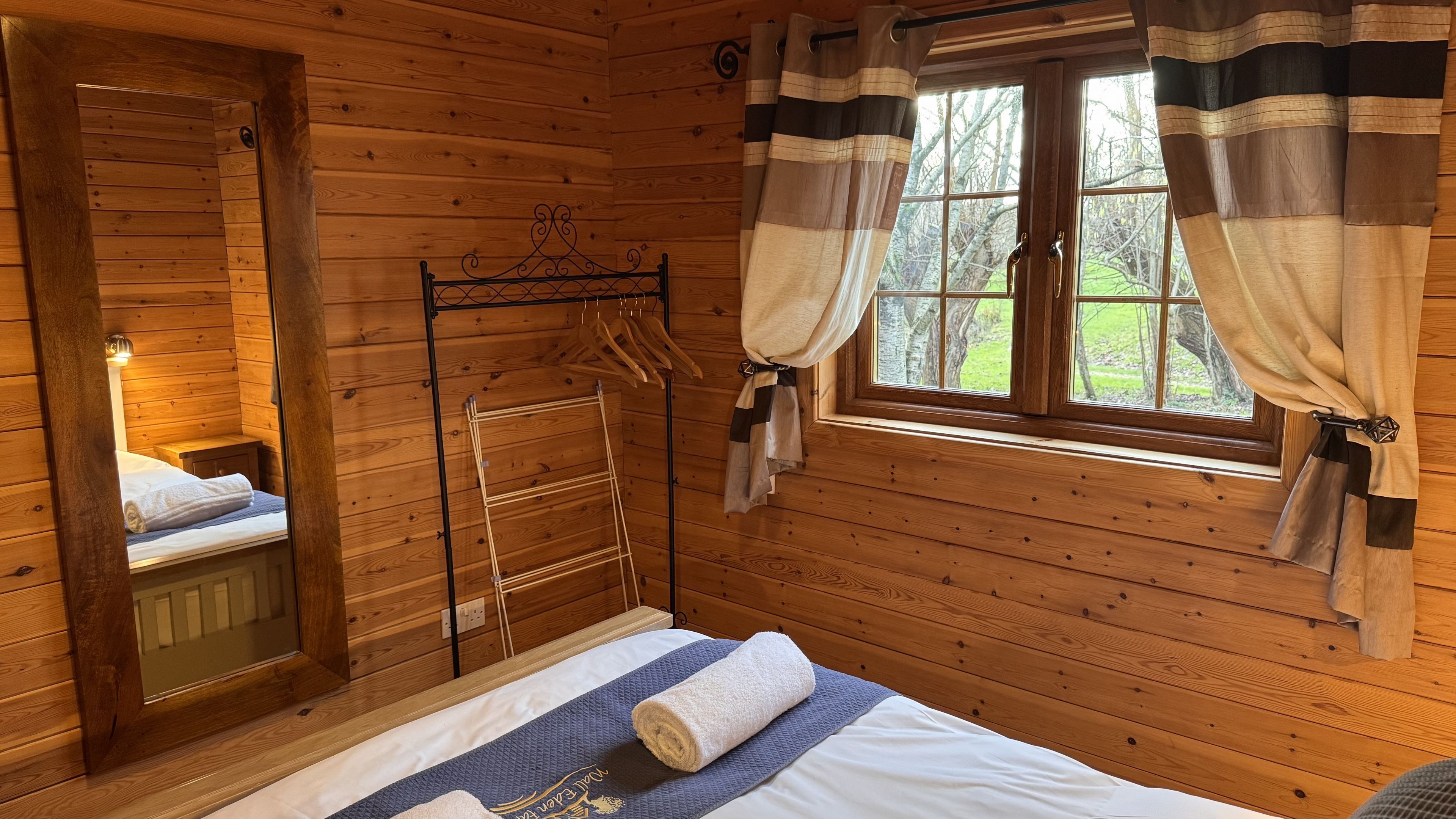 A wooden cabin room with a bed, a hanging clothes rack, a mirror, and a window with curtains.
