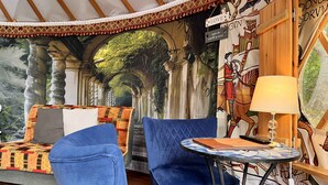 Een gezellige kamer met een blauwe bank, een klein rond tafeltje met een lamp, en een muurschildering van een bosrijk pad.