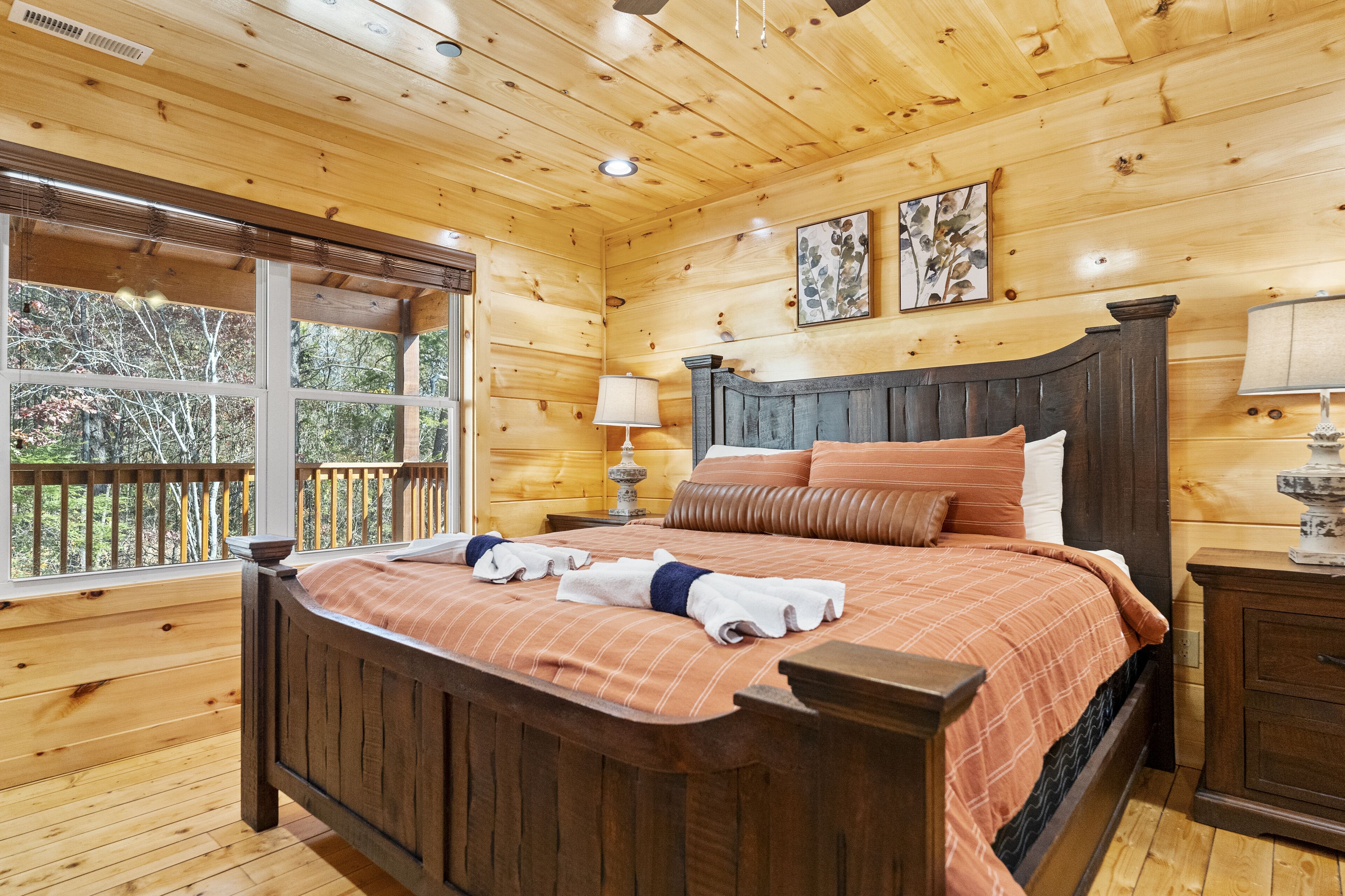 A wooden cabin bedroom with a large bed, a nightstand, a lamp, and a window overlooking a deck.