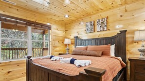 Ein Schlafzimmer einer Holzhütte mit einem großen Bett, einem Nachttisch, einer Lampe und einem Fenster mit Blick auf eine Terrasse.
