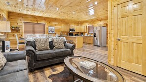 Ein rustikales Blockhaus-Interieur mit einem Wohnbereich, der ein Sofa und einen Glastisch umfasst, einer Küche mit Holzausstattung und Geräten sowie einer großen Holztür.