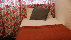 Una cama pequeña y ordenada con una colcha roja y dos almohadas, una blanca y otra gris, en una habitación con cortinas estampadas.
