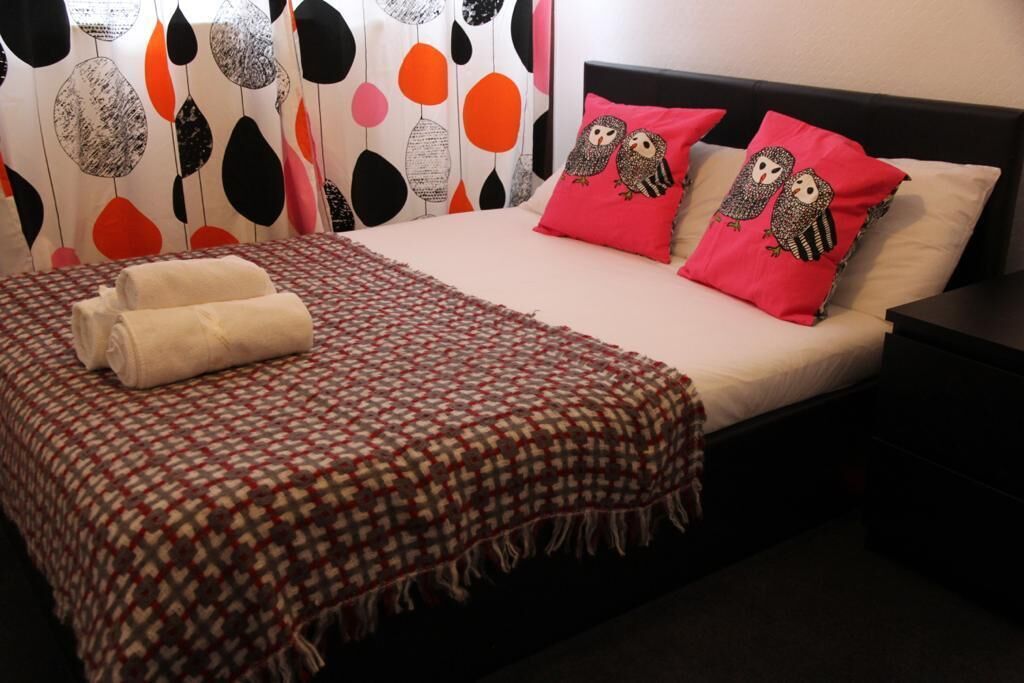 A neatly made bed with a checkered bedspread, two red pillows with owl designs, and a rolled white towel.