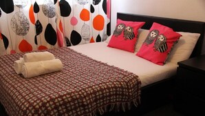 Una cama bien hecha con una colcha a cuadros, dos almohadas rojas con diseños de búhos y una toalla blanca enrollada.