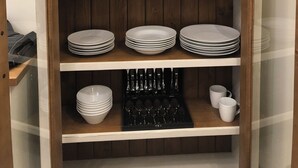 Un mueble de madera con estantes que contiene platos blancos, tazas, copas de vino y cubiertos.