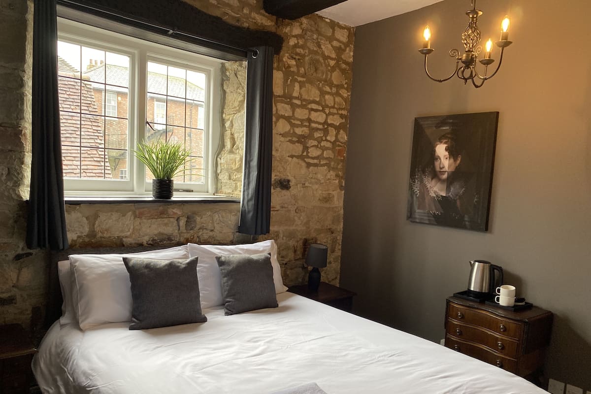 A bedroom with a large bed, a window with curtains, a potted plant, a painting, a nightstand, a lamp, and a dresser.