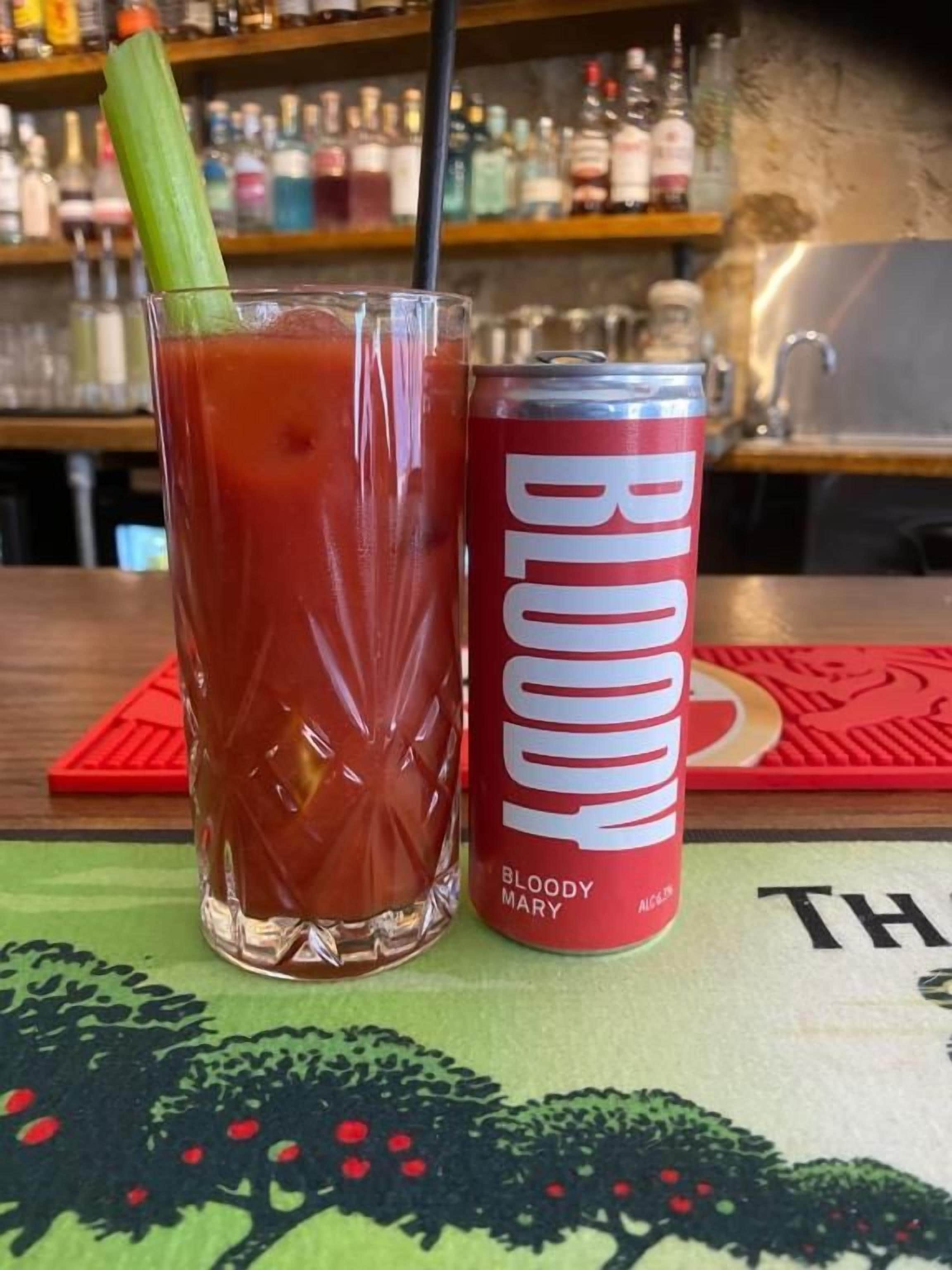 A Bloody Mary cocktail in a glass with a celery stalk, next to a can of BUD light beer on a bar counter.