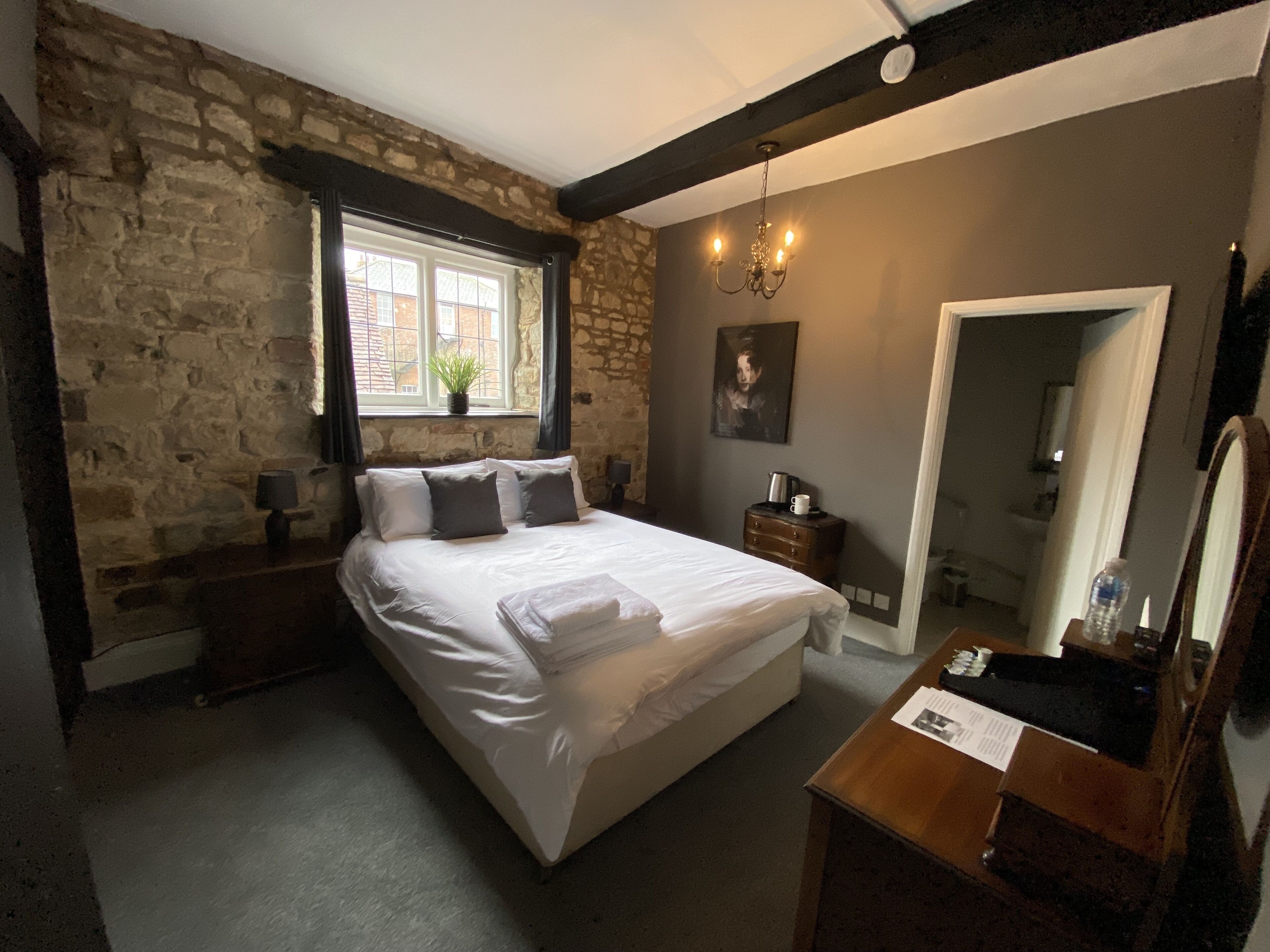 A bedroom with a stone wall, a large bed, a window with curtains, a bedside table with a lamp, a dresser, a mirror, and a door.
