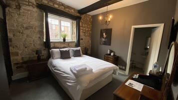 A bedroom with a stone wall, a large bed, a window with curtains, a bedside table with a lamp, a dresser, a mirror, and a door.