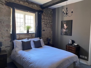 A bedroom with a stone wall, a large bed with white and dark blue bedding, a potted plant by the window, a painting on the wall, and a bedside table with a lamp. - The Bull (Battle)