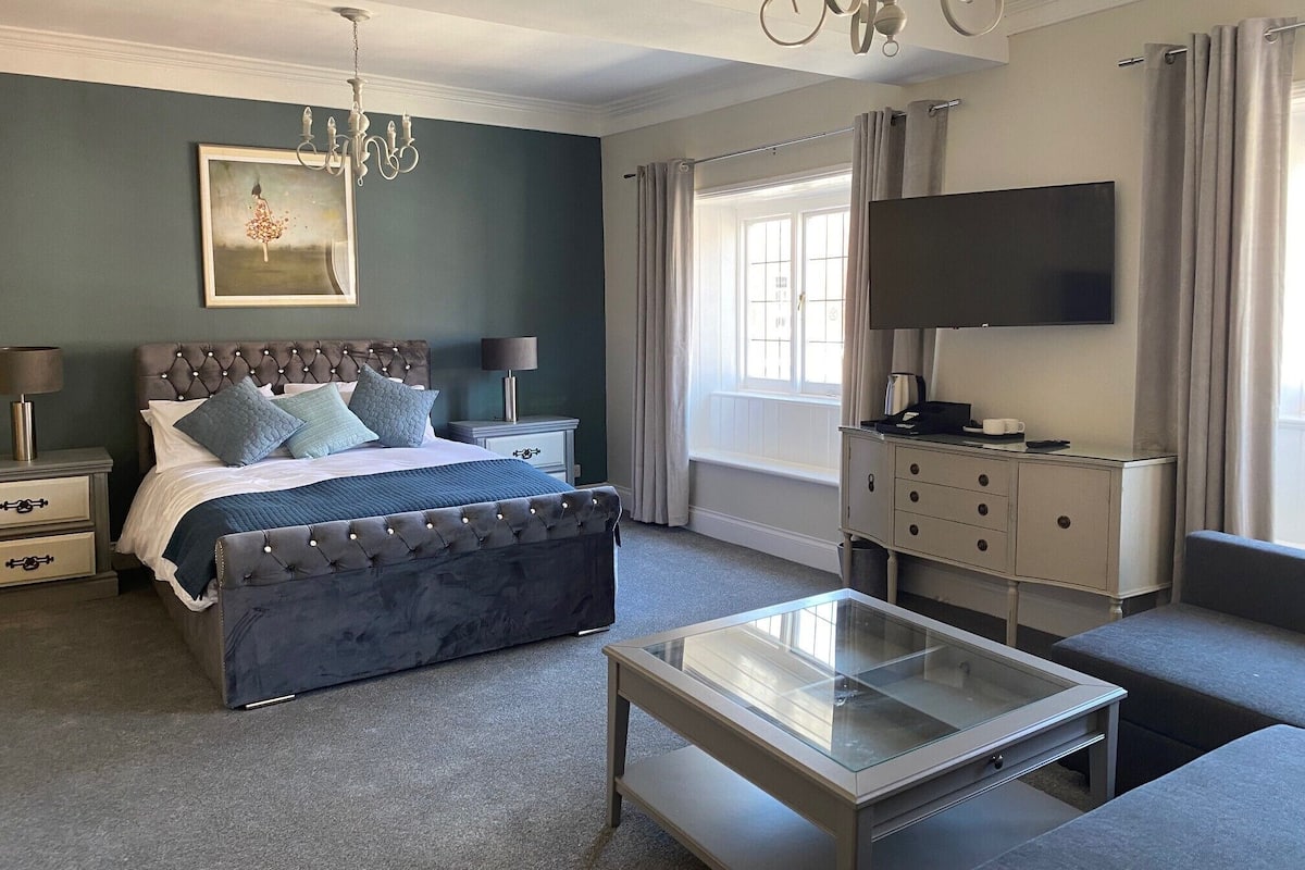 A bedroom with a tufted headboard bed, a glass coffee table, a television, and a chandelier.