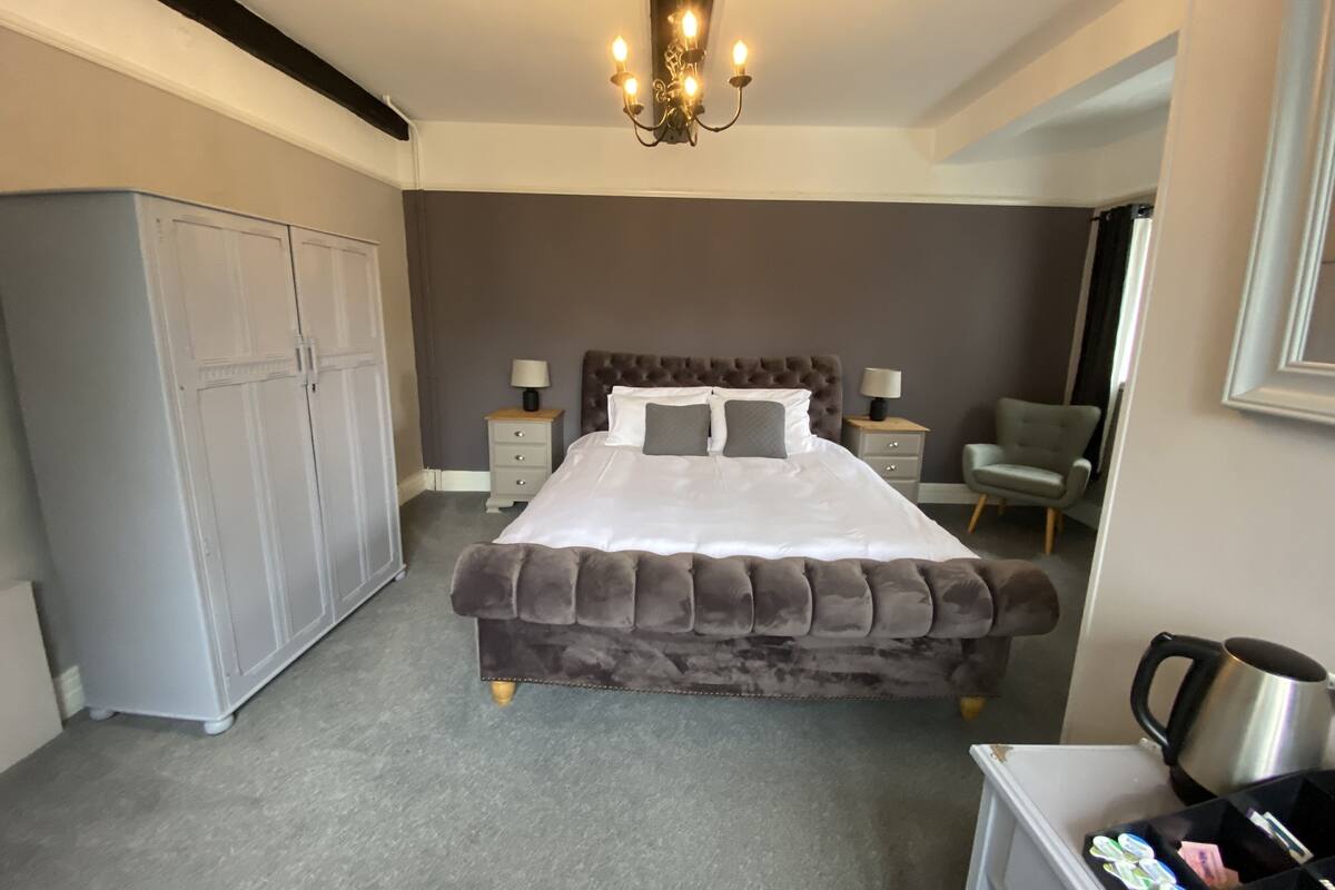 A bedroom with a large bed, two bedside tables, a wardrobe, a chair, and a coffee maker.