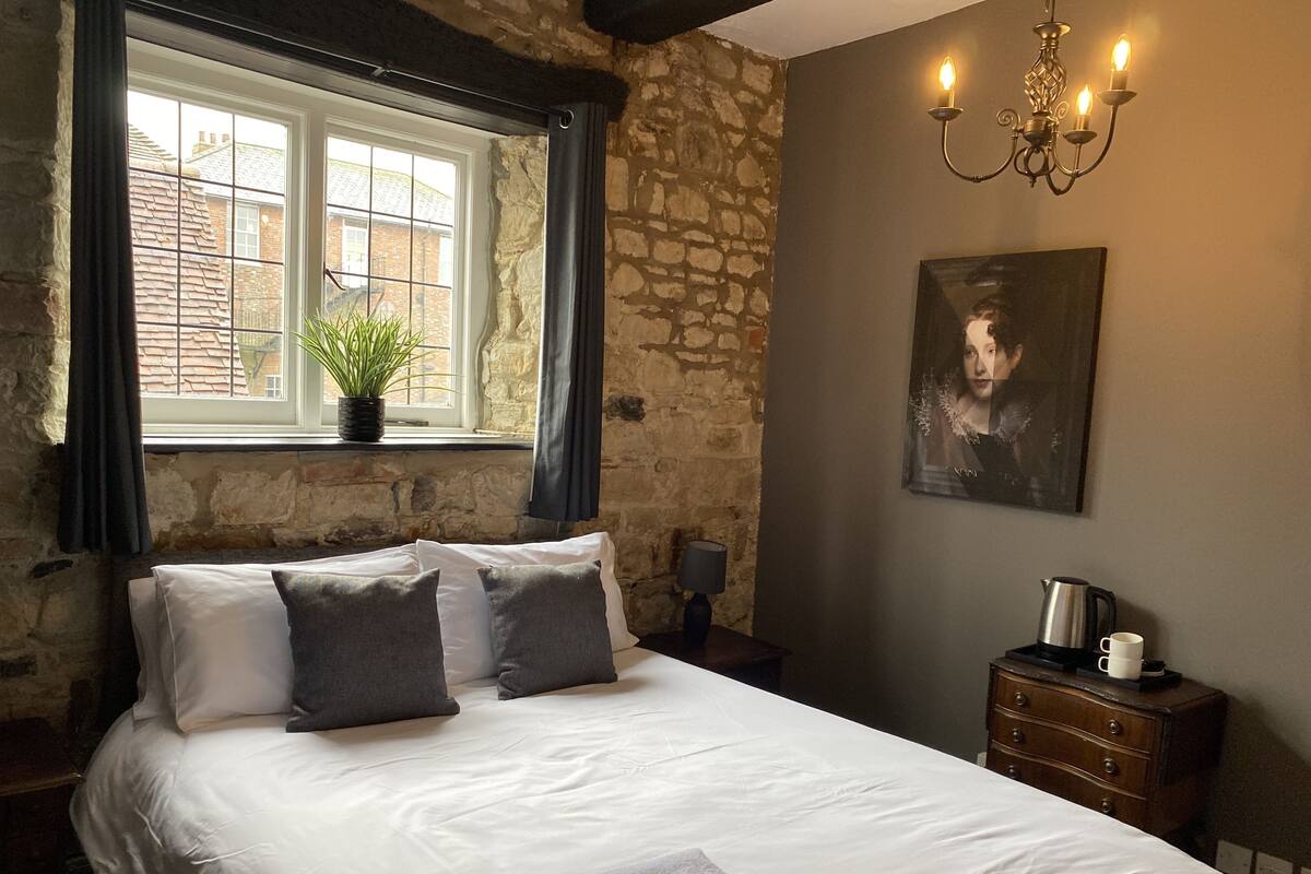 A bedroom with a bed, a window with curtains, a potted plant, a painting, a nightstand, a lamp, a dresser, and a kettle.