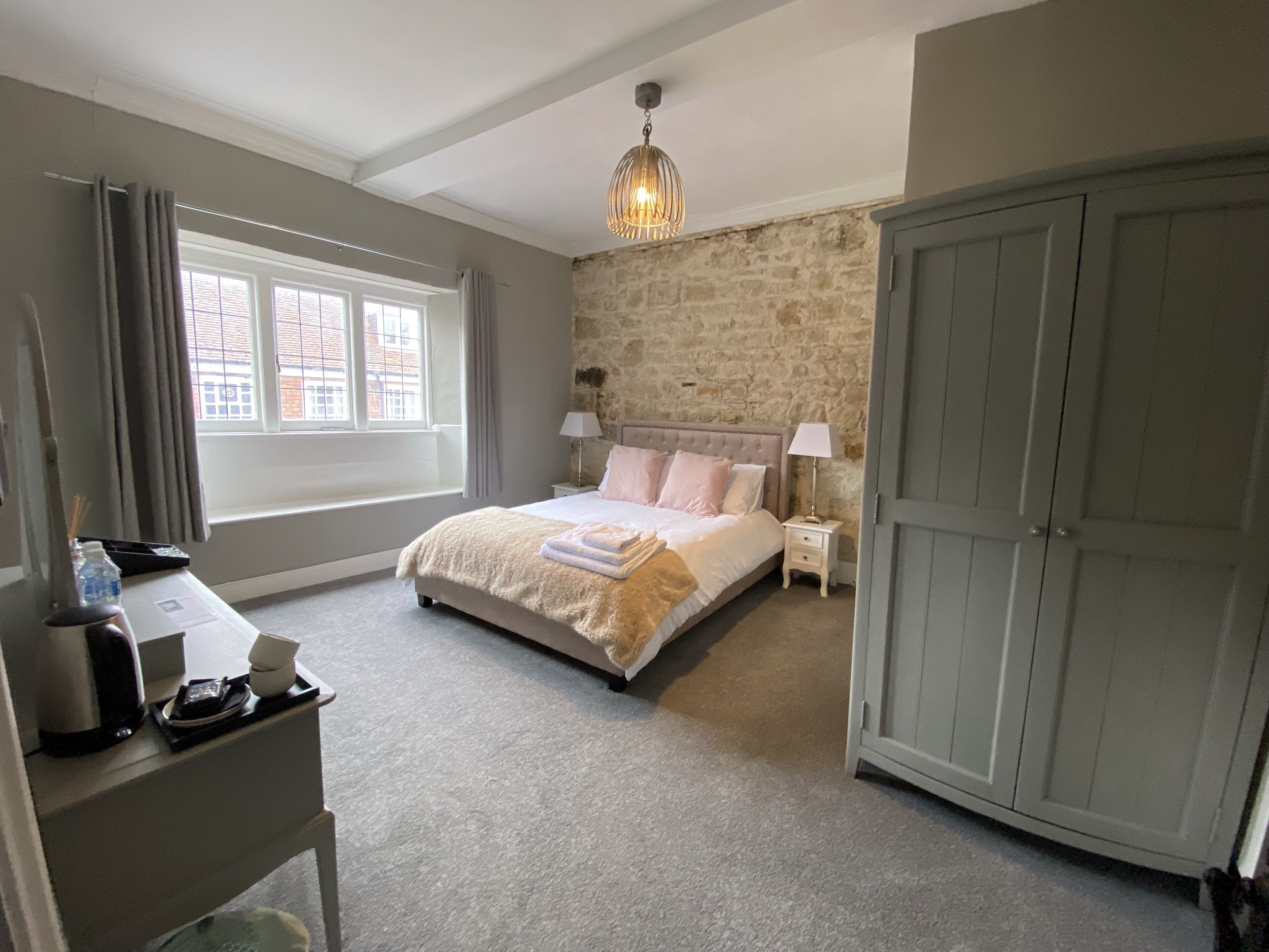 A bedroom with a stone wall, a bed with a beige and pink bedspread, a nightstand with a lamp, a wardrobe, and a desk with a coffee maker.