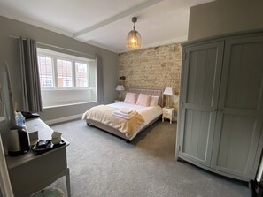 A bedroom with a stone wall, a bed with a beige and pink bedspread, a nightstand with a lamp, a wardrobe, and a desk with a coffee maker. - The Bull (Battle)