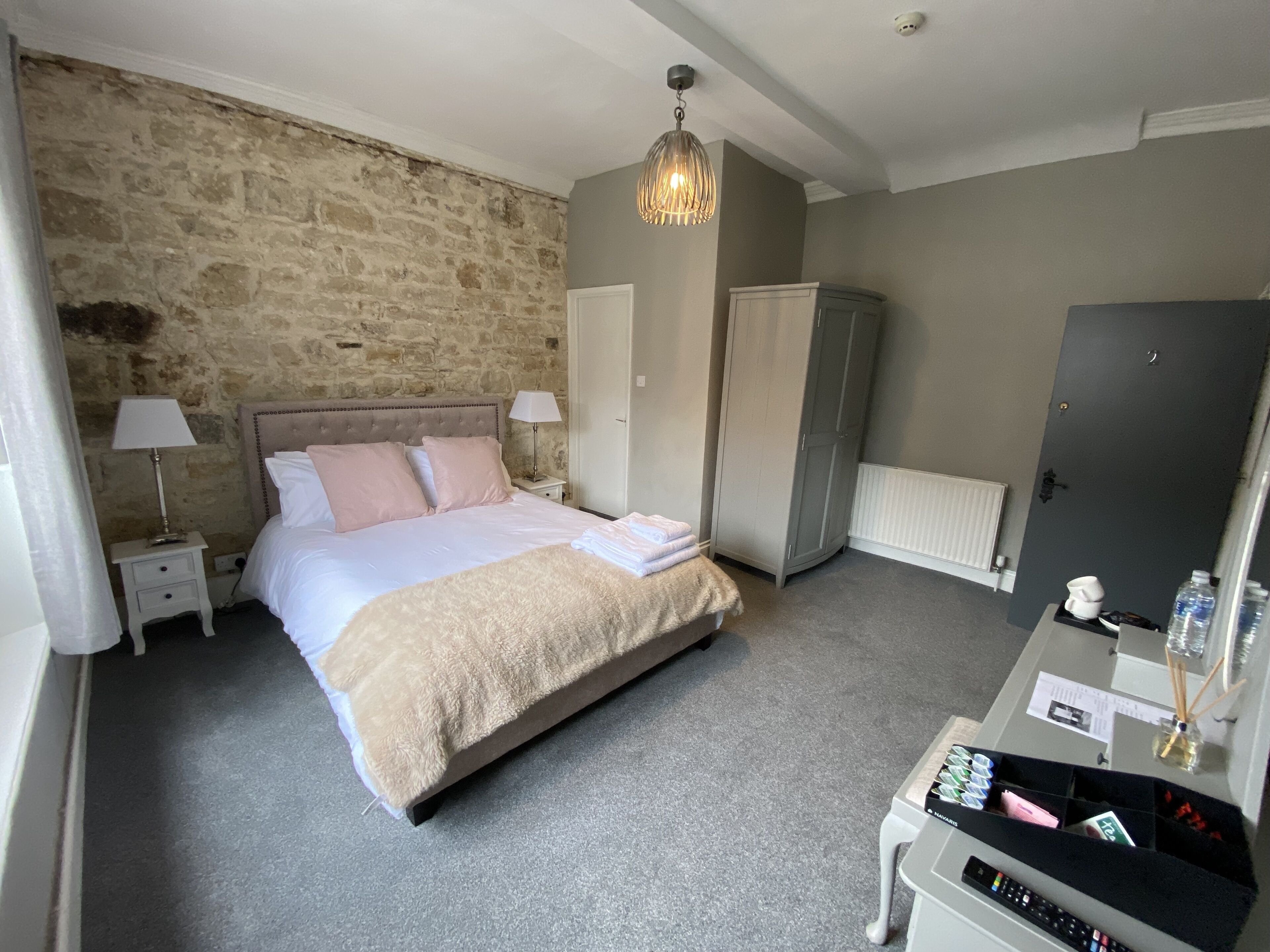 A bedroom with a stone wall, a bed with a beige blanket, a wardrobe, a desk with a computer, and a lamp.