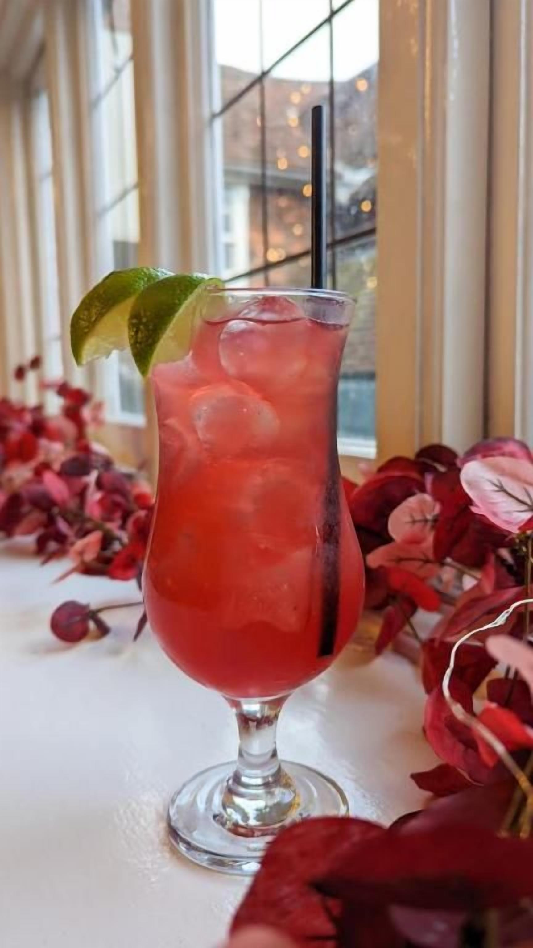 Un vaso de bebida roja con un gajo de lima y cubitos de hielo, sobre una mesa con flores rojas.