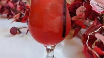Un vaso de bebida roja con un gajo de lima y cubitos de hielo, sobre una mesa con flores rojas.