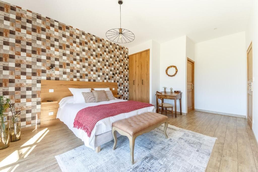 Une chambre avec un mur à carreaux, une tête de lit en bois, un lit recouvert d’une couverture rouge, un banc, une petite table avec des chaises et un luminaire suspendu.