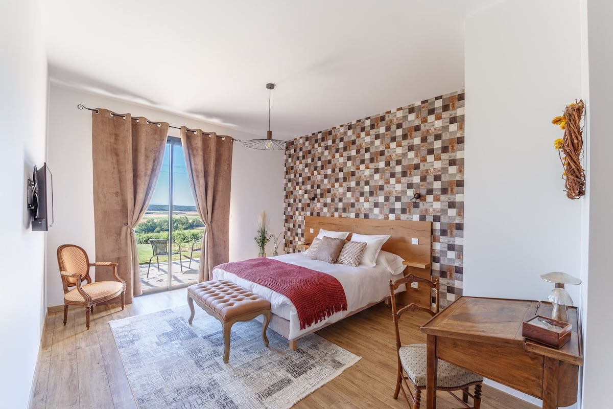 Quarto com uma cama grande, cabeceira de madeira, vista para o exterior, uma mesa pequena e uma cadeira.