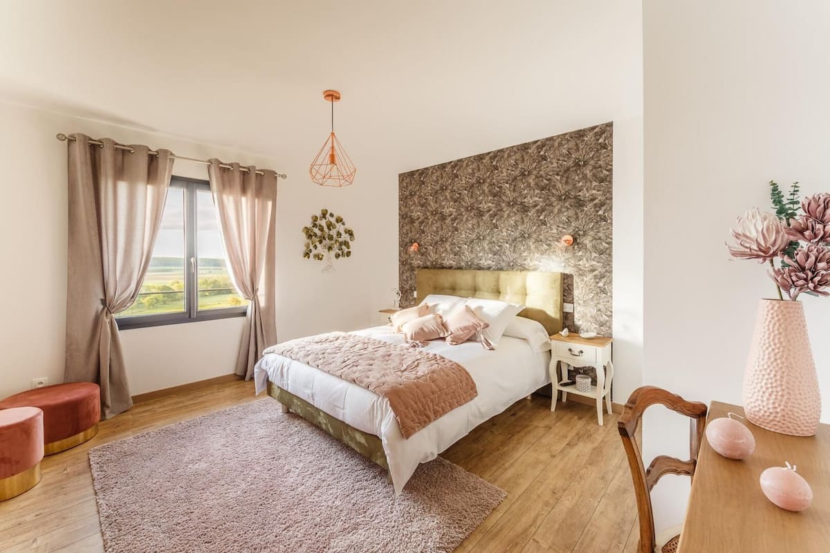 Quarto com uma cama grande, mesa de cabeceira, cadeira, mesa de madeira e janela com cortinas.