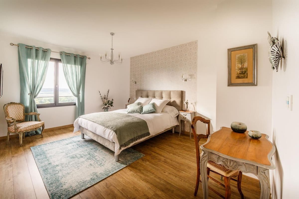 Quarto com uma cama grande, uma mesa de madeira, uma cadeira, uma mesa de cabeceira, um candelabro e uma janela com cortinas.