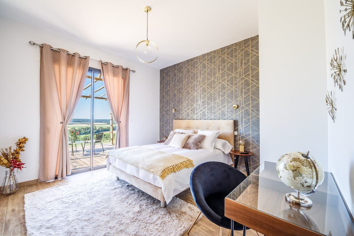 Quarto com cama, escrivaninha com globo, cadeira, janela com cortinas e parede estampada.