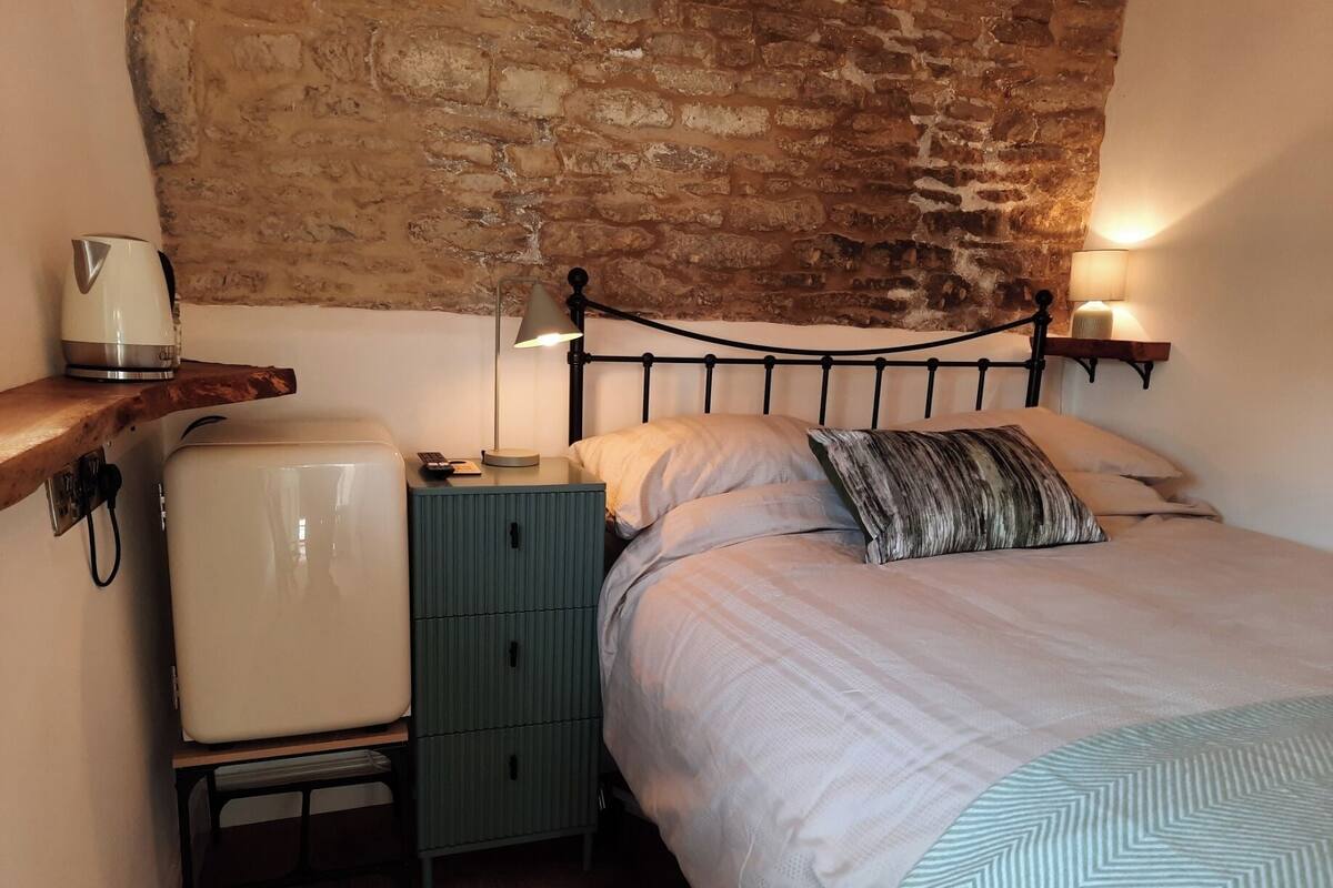A bedroom with a brick wall, a bed with a patterned blanket, a bedside table with a lamp, and a small refrigerator.