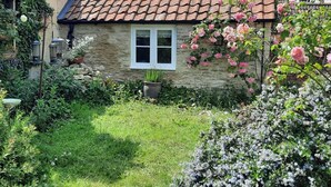 Uma casa de pedra com telhado de telhas, uma pequena janela e um jardim com diversas plantas e flores.