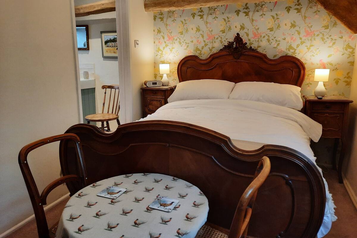 A traditional bedroom with a wooden bed, two bedside tables with lamps, a round table with a bird pattern, and floral wallpaper.