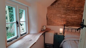 Quarto pequeno e aconchegante com um banco de madeira junto à janela, cama com cabeceira, mesa de cabeceira com abajur e um pequeno frigorífico.