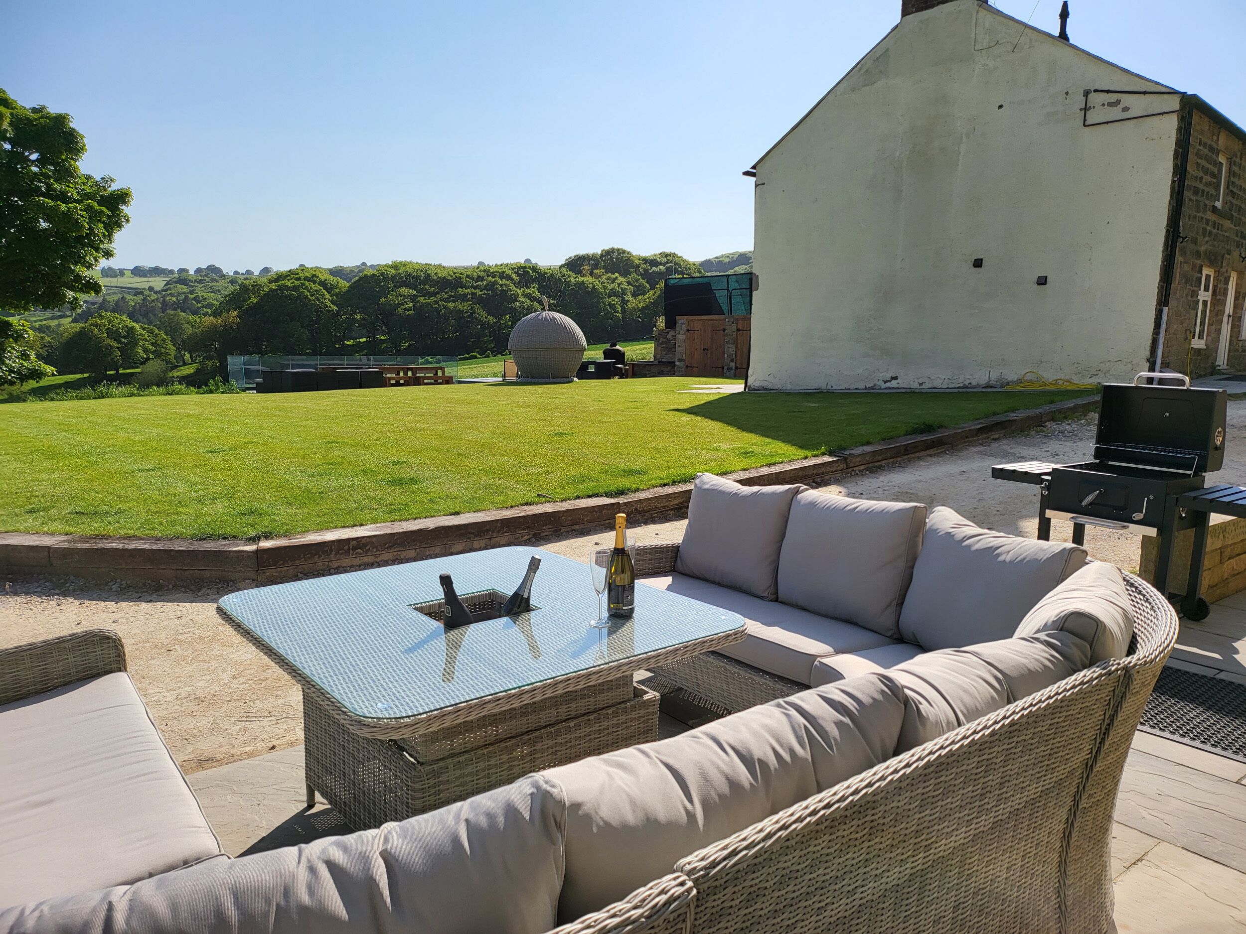 Un área de estar al aire libre con una mesa de vidrio y muebles de mimbre, una parrilla y vistas a un campo de césped y un edificio grande al fondo.