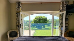 Un dormitorio con una cama grande, vistas a un paisaje verde a través de un ventanal, y mobiliario de mimbre.