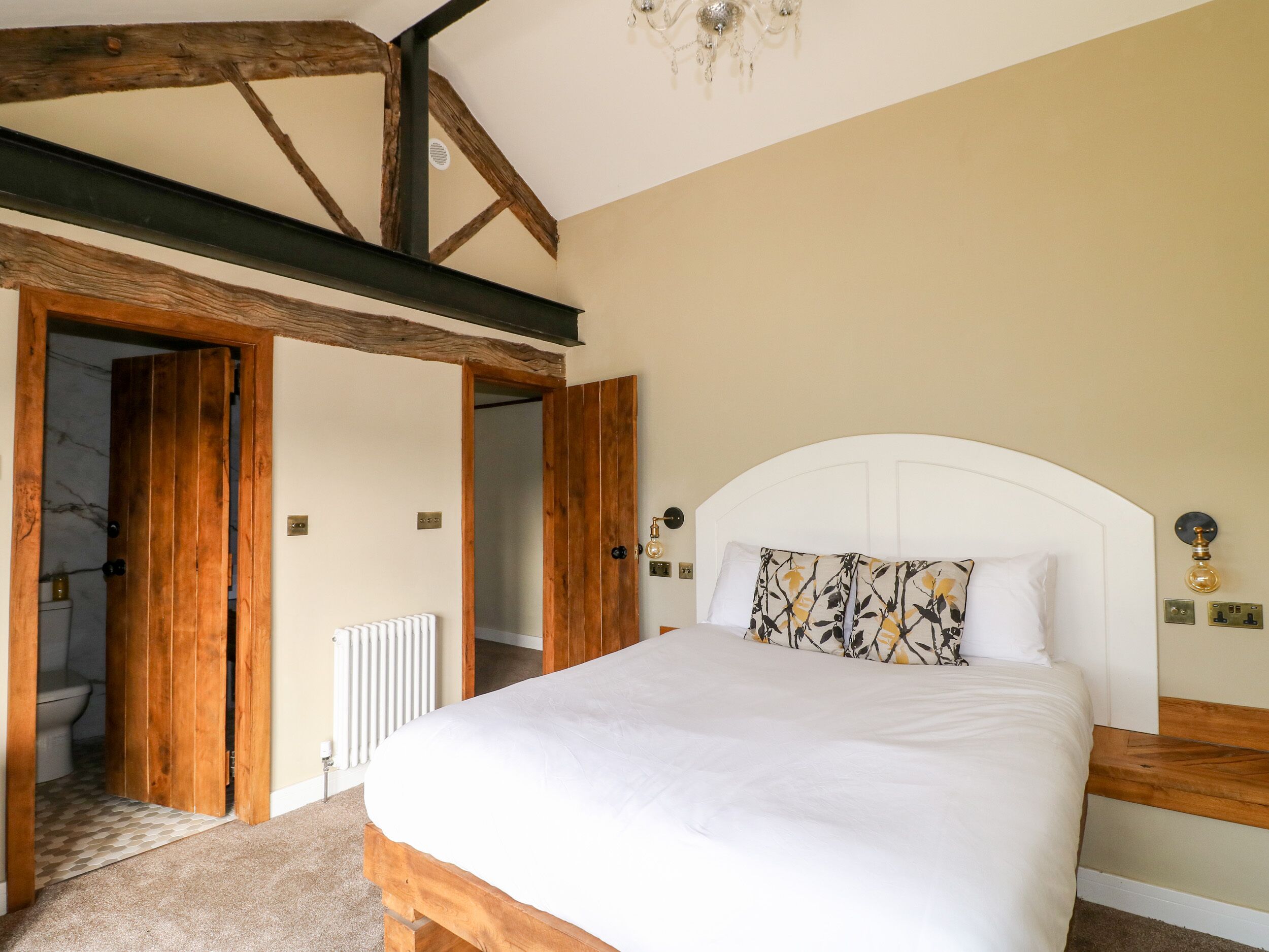 A bedroom with a wooden headboard, a white bedspread, and a patterned pillow. There is a radiator on the wall and a wooden door leading to another room.