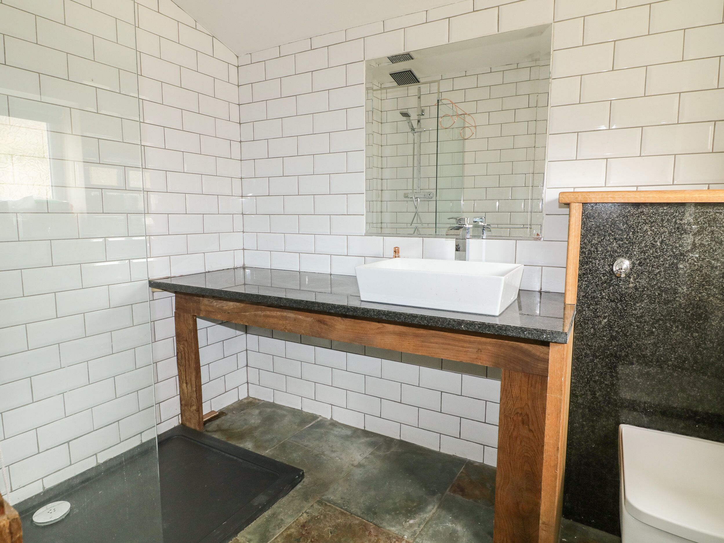 A bathroom with white tiled walls, a wooden vanity, a white sink, and a black countertop.
