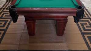 A pool table with a green felt surface, surrounded by a patterned rug, and a view of a wooded area through a glass door. - Southern Comfort Cabin- minutes from the river and the lake! (Murfreesboro)