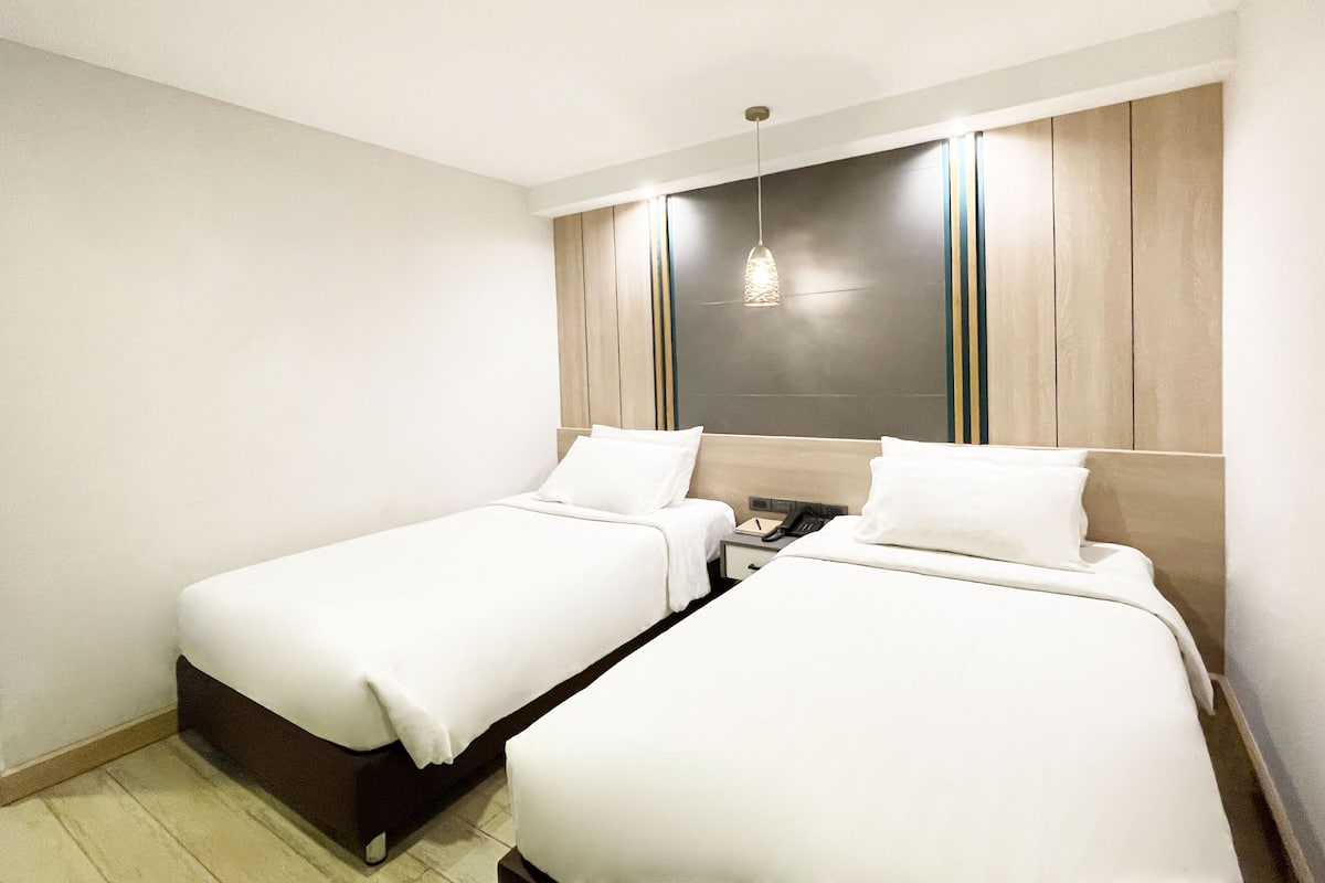 A hotel room with two single beds, a wooden headboard, a bedside table with a lamp, and a small nightstand.