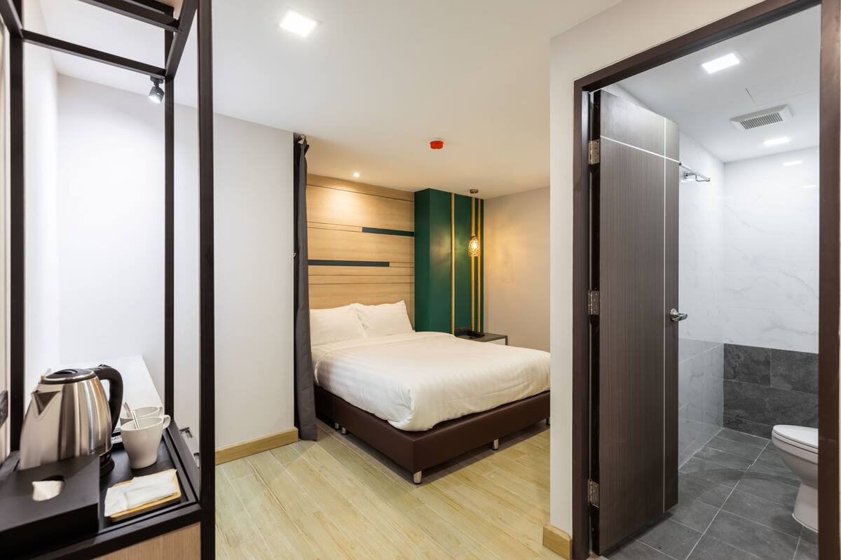 A modern hotel room with a bed, a bedside table with a kettle and cups, a shower area, and a door leading to another room.