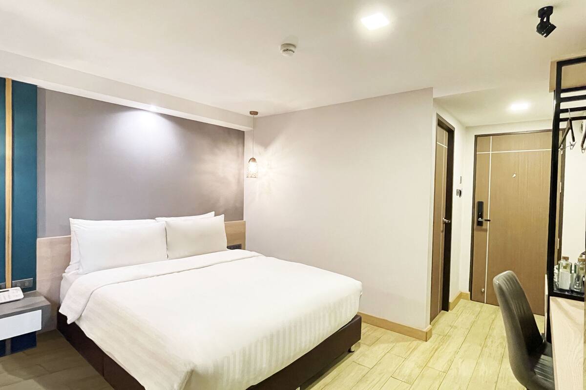 A hotel room with a large bed, a desk with a chair, a wardrobe, and a door.