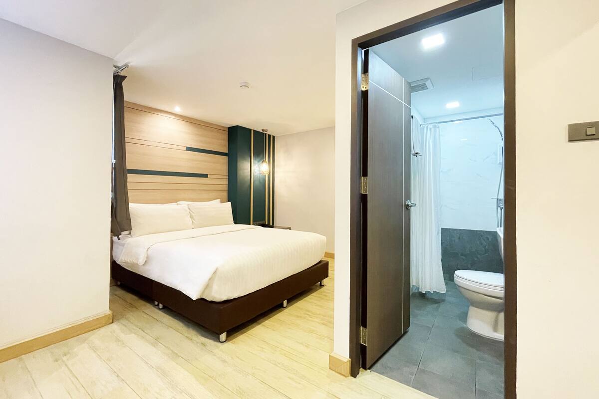 A modern bedroom with a bed, a wooden headboard, and a view into the bathroom through an open door.